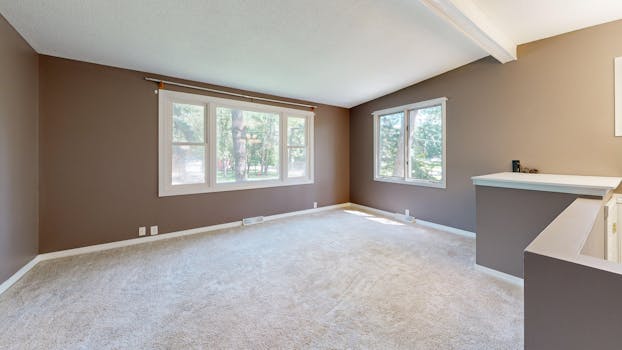 Large empty living room with neutral walls and carpet, natural light through windows.