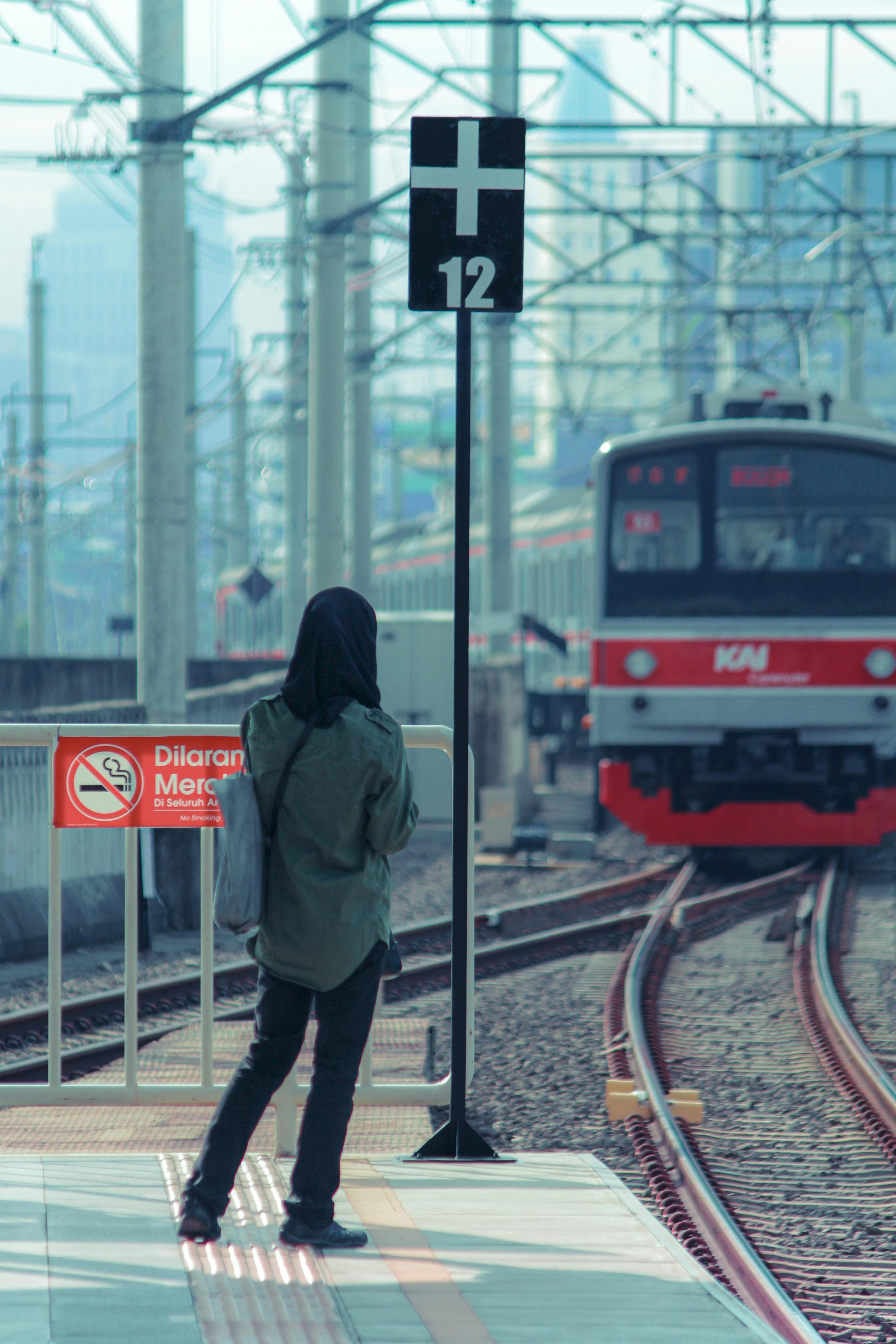 A person in a hooded jacket stands on a train platform as a train approaches on a sunny day.
