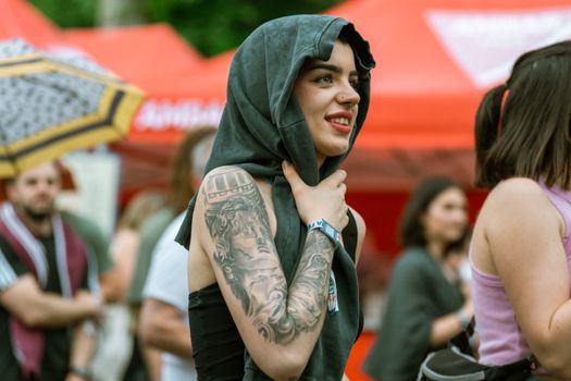 Smiling woman with tattoo and hood at a vibrant outdoor summer festival.