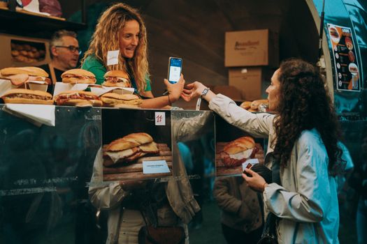 Two women exchanging digital payment at a food truck selling sandwiches outdoors in a bustling setting.