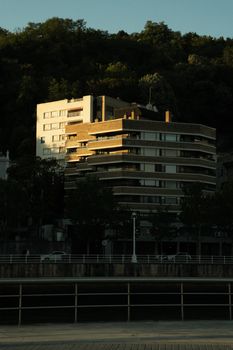 A contemporary urban building by the river during sunset, casting dramatic shadows.