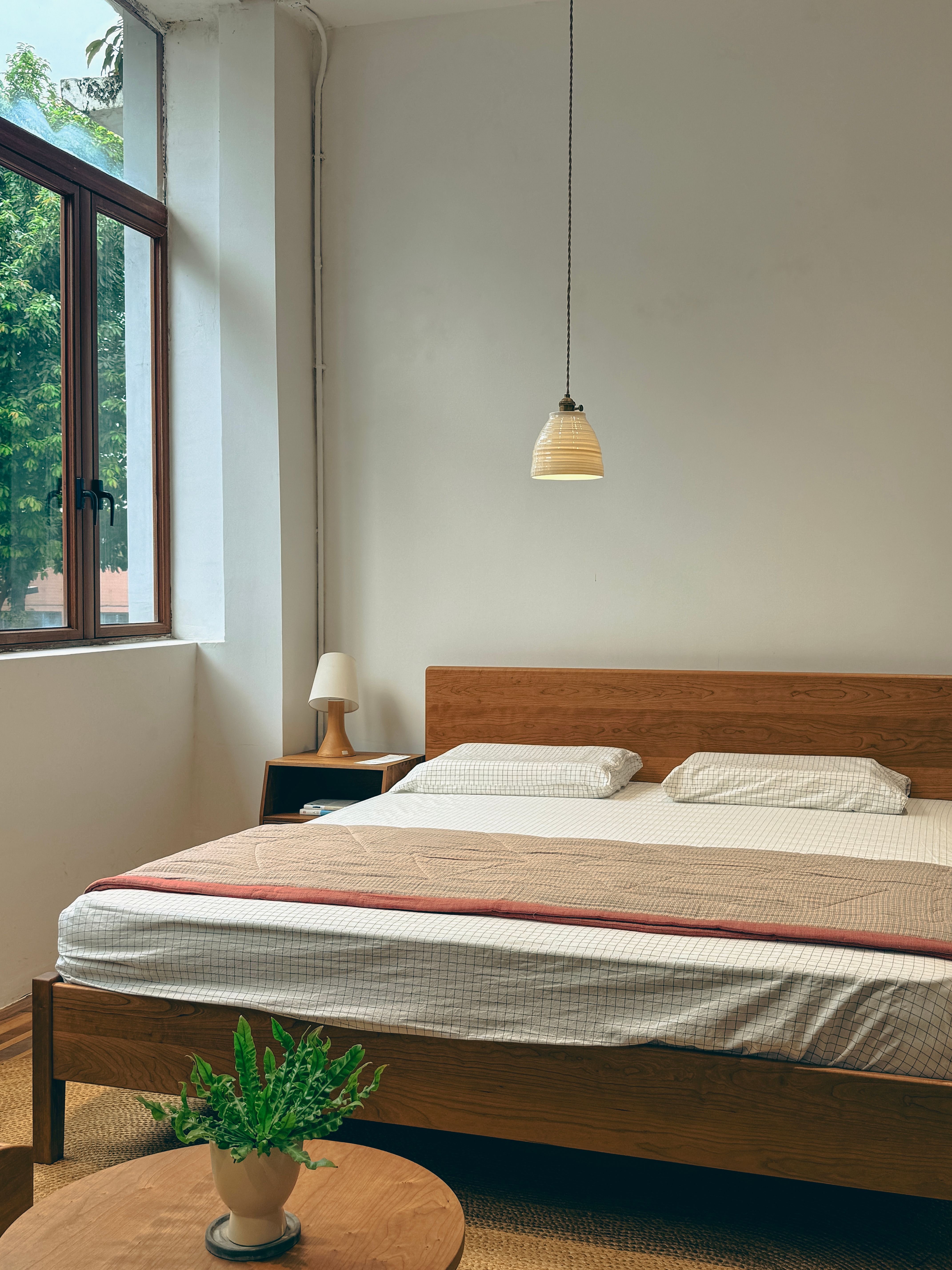 Minimalist bedroom with a low wooden bed, white walls, single pendant, large window and one small plant