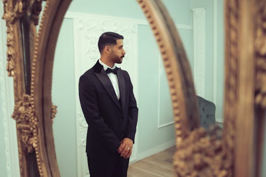 A sophisticated man in a tuxedo stands by an ornate mirror in an elegant room.