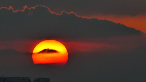 A breathtaking view of the sun setting behind dark clouds with vibrant red hues dominating the sky.