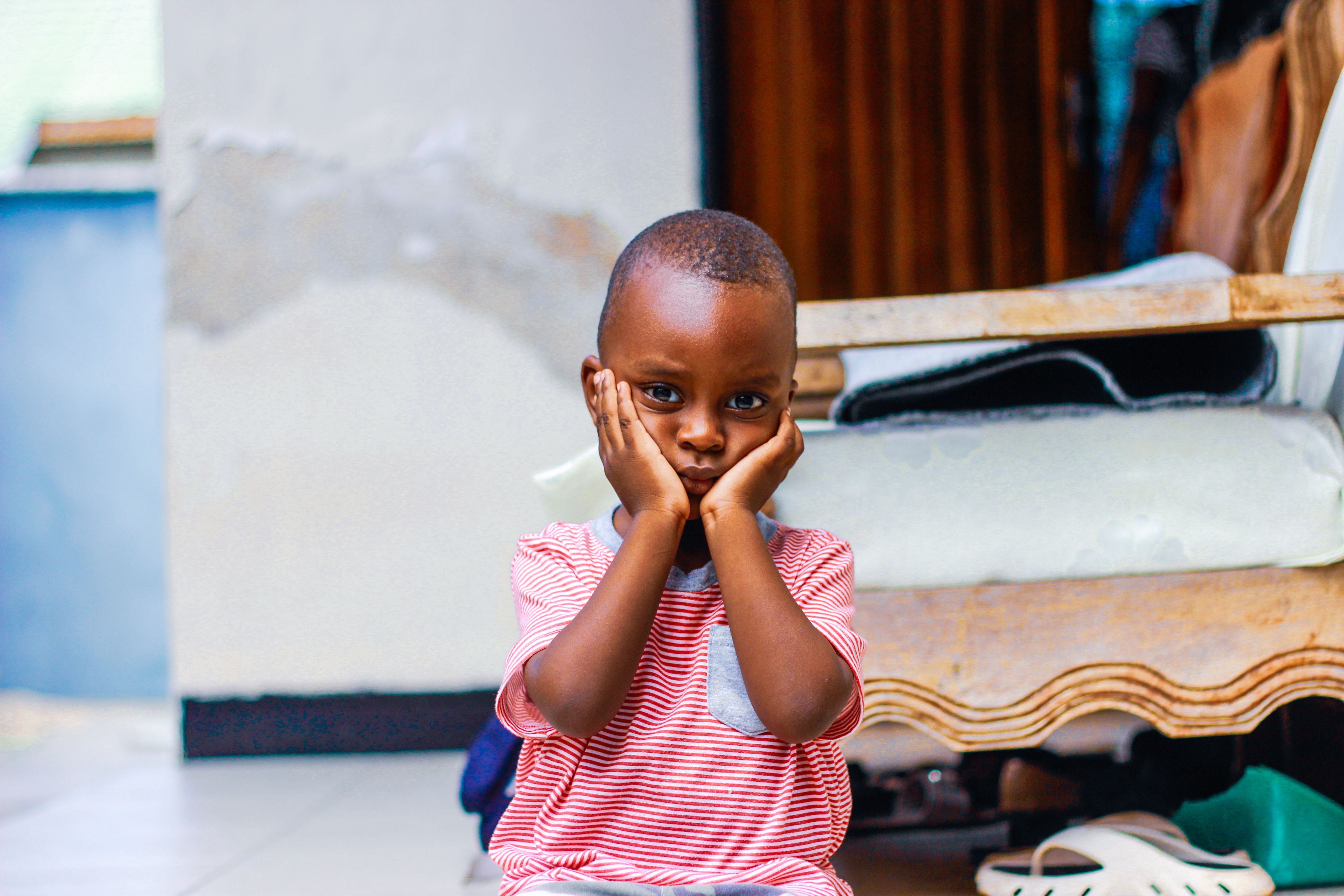 A young boy in a striped shirt sits on a porch, hands on his cheeks, looking thoughtful.