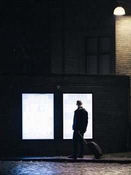 A person stands at night looking at illuminated posters on an urban wall with a suitcase.