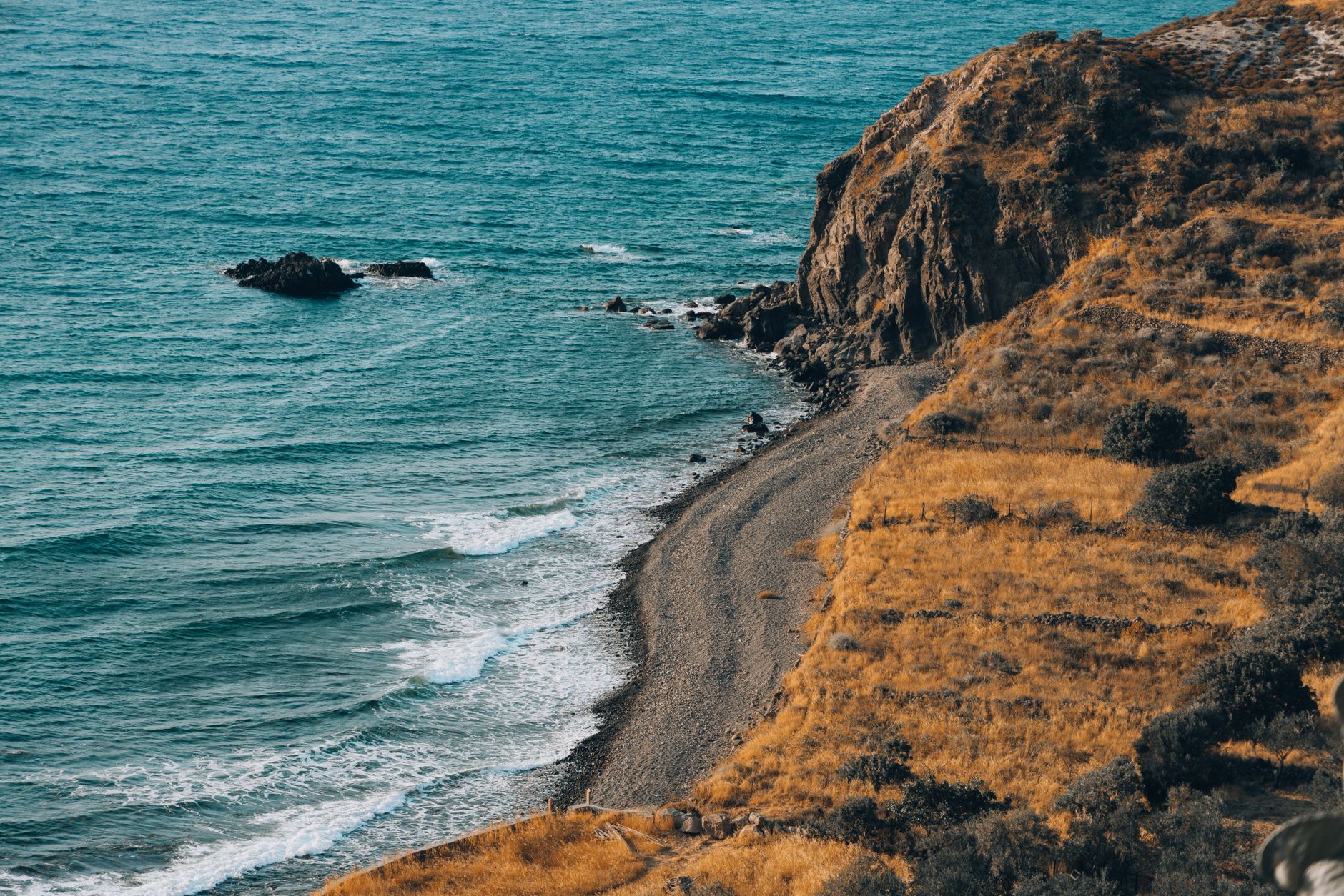 Dramatic aerial shot of a rugged coastal cliff and pebble beach with turquoise ocean waves.