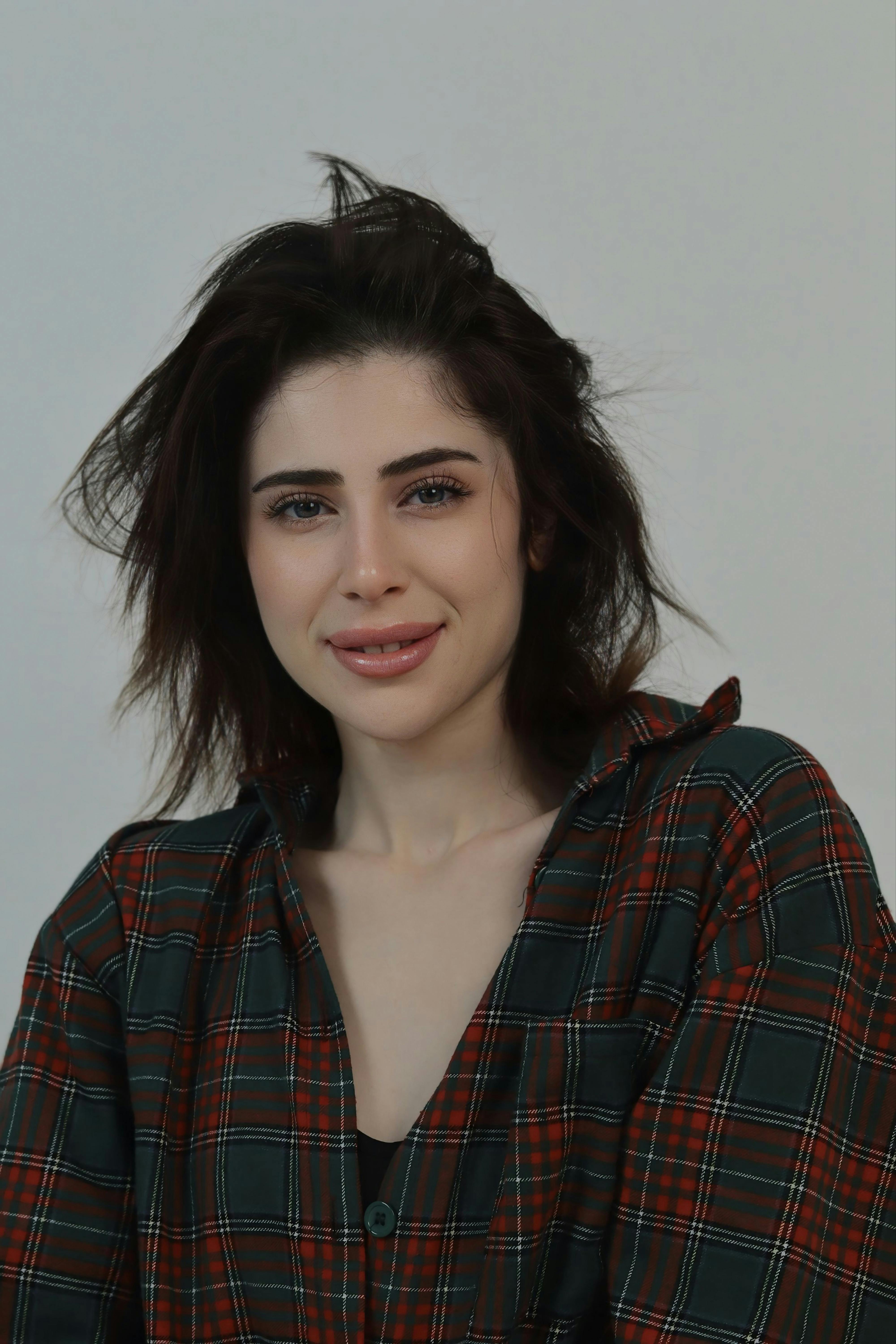 Kostenlos Frau in kariertem Hemd lächelt sanft vor einem schlichten Hintergrund und vermittelt Gemütlichkeit und Stil. Stock-Foto