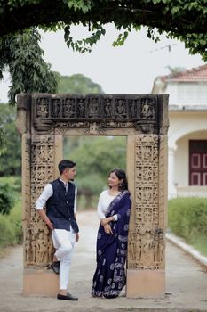 A couple poses in traditional Indian clothing beside an ancient stone arch outdoors.