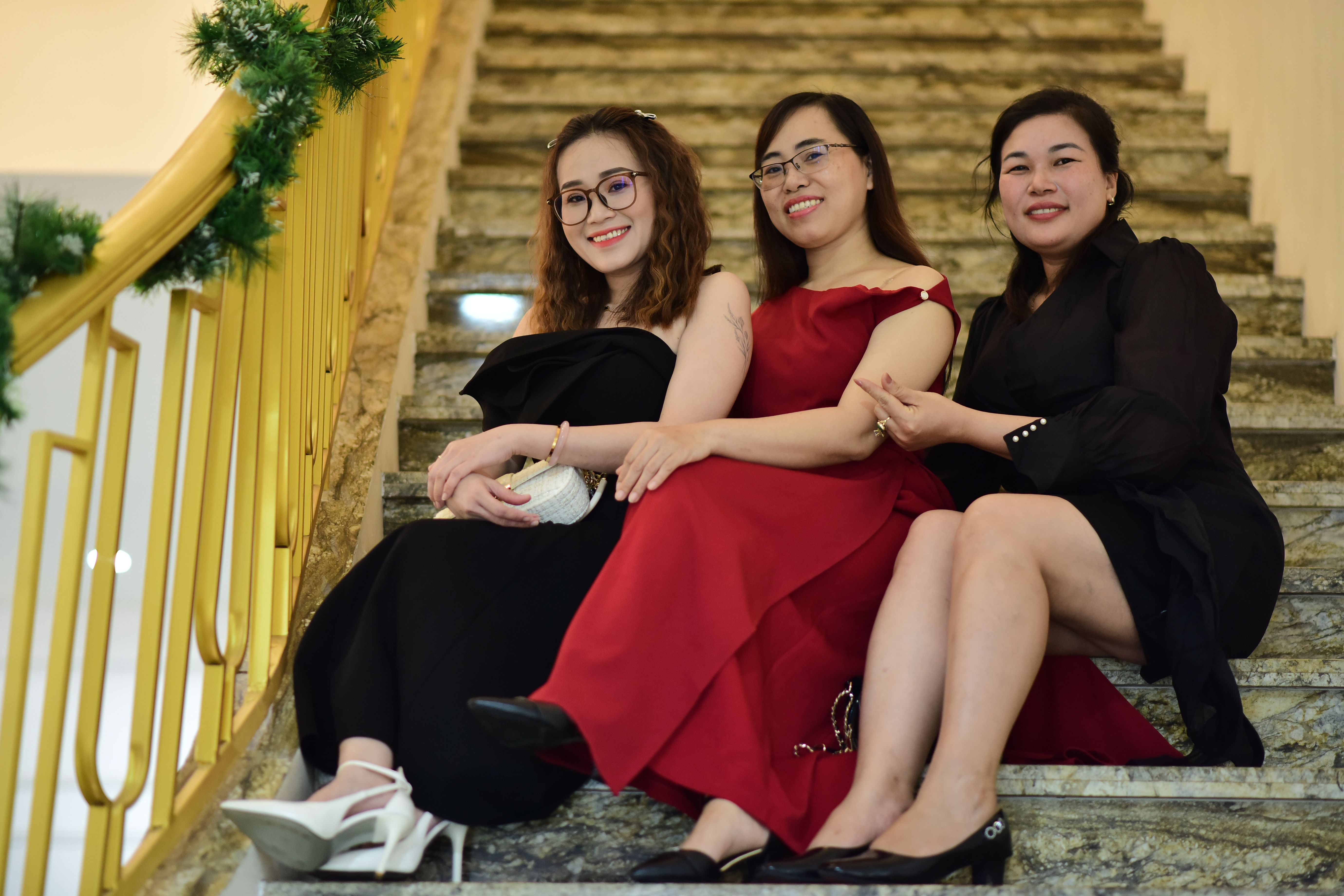 Three women dressed stylishly pose on a decorated staircase indoors.