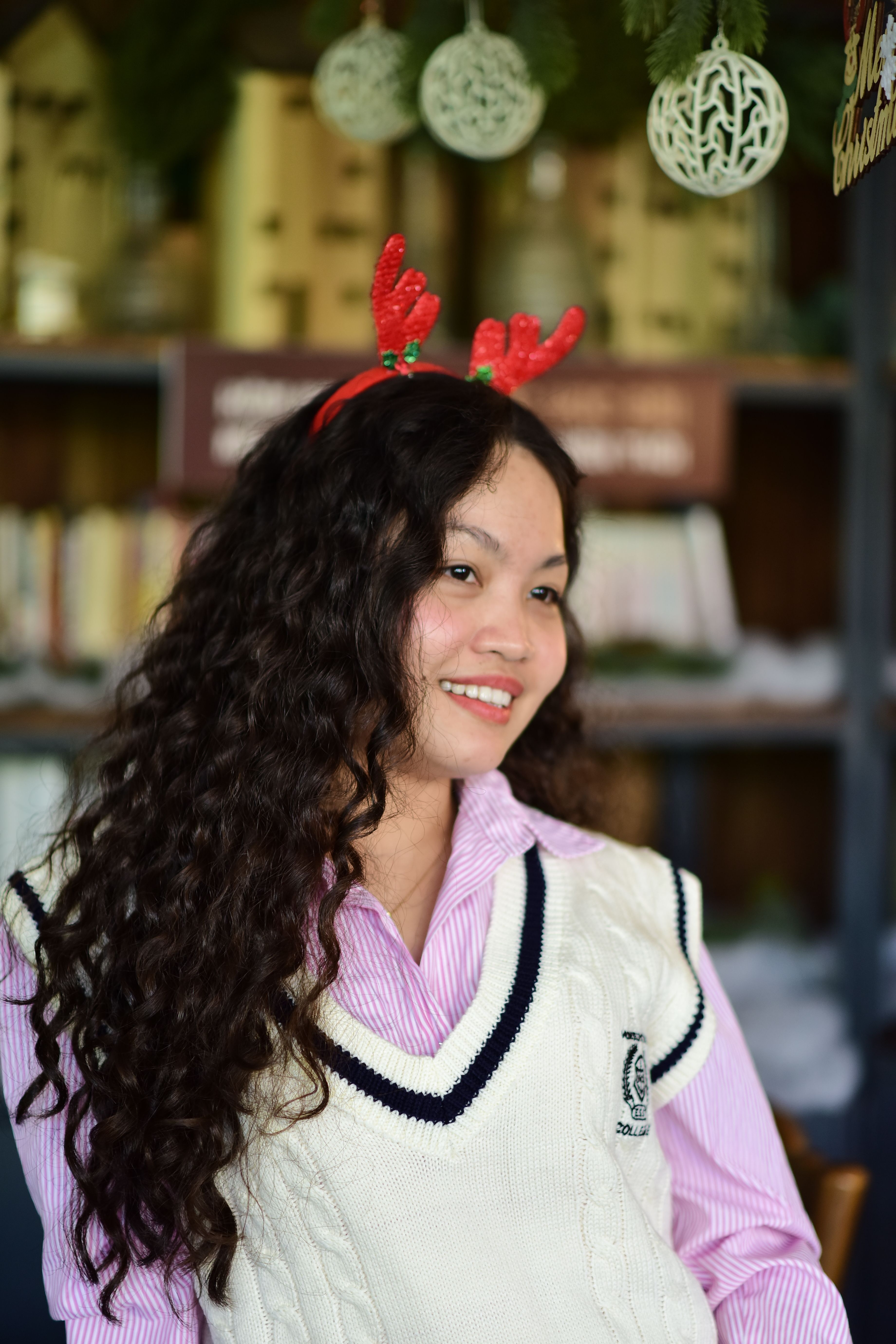 Happy woman with curly hair wearing reindeer antlers headband, enjoying festive indoor setting.