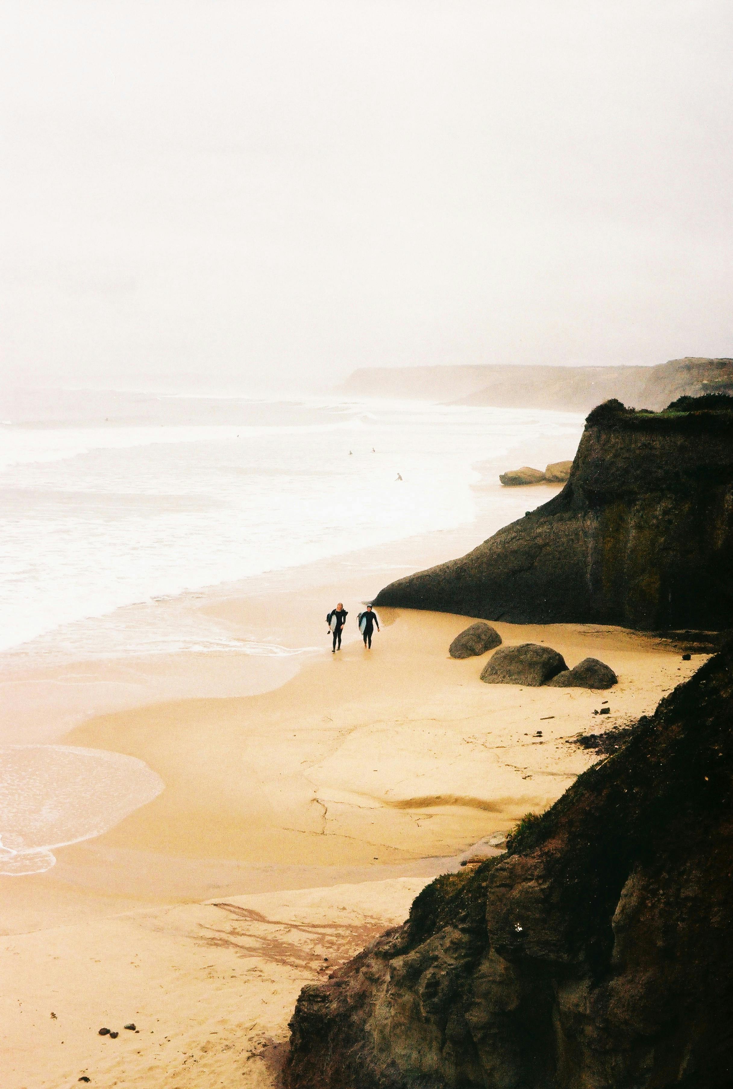 Two surfers walk along a misty beach with towering cliffs and crashing waves.