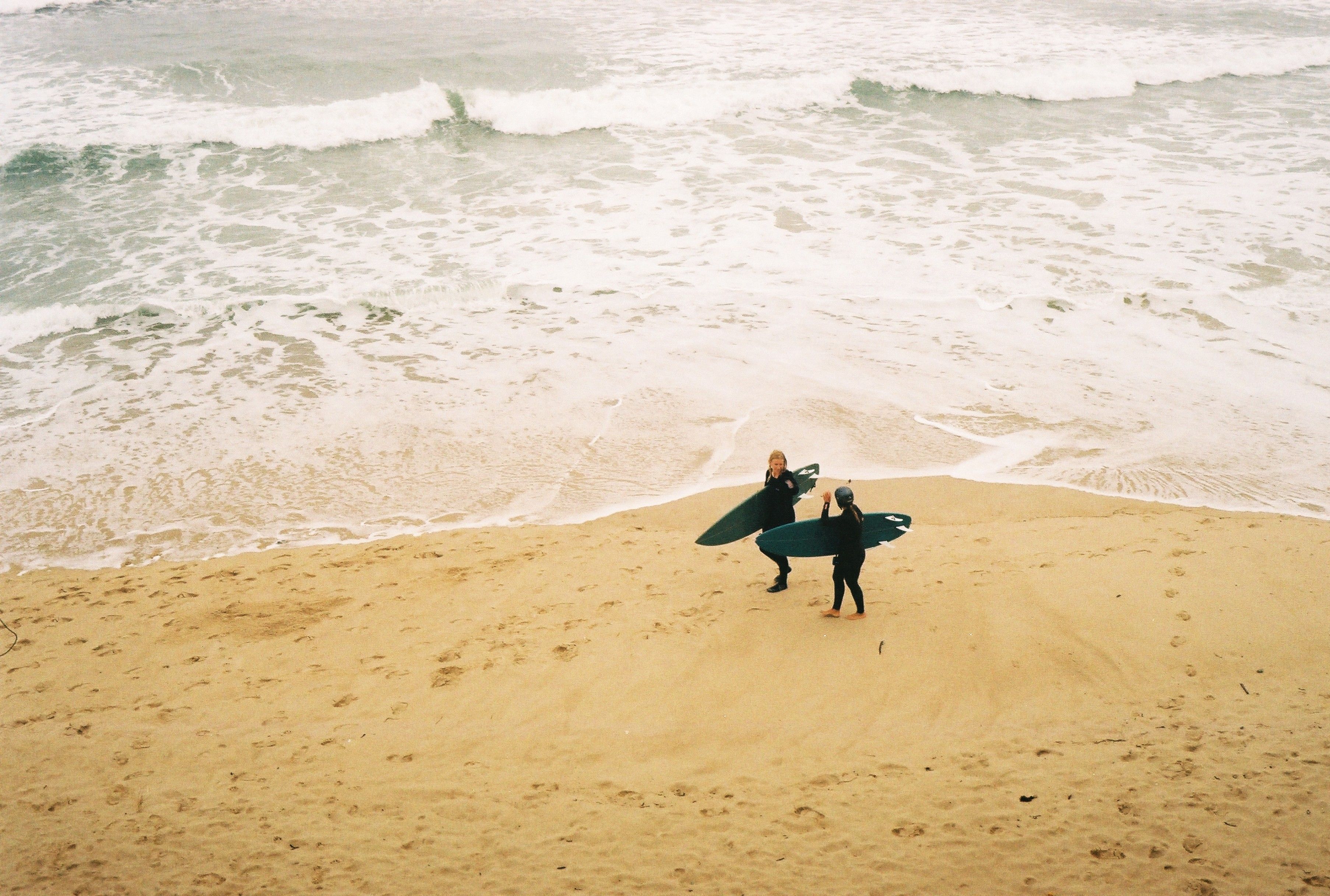 Two surfers with boards on a sandy beach prepare to surf in the ocean waves.