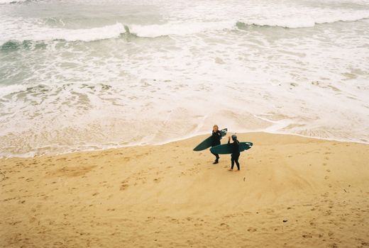 Two surfers with boards on a sandy beach prepare to surf in the ocean waves.