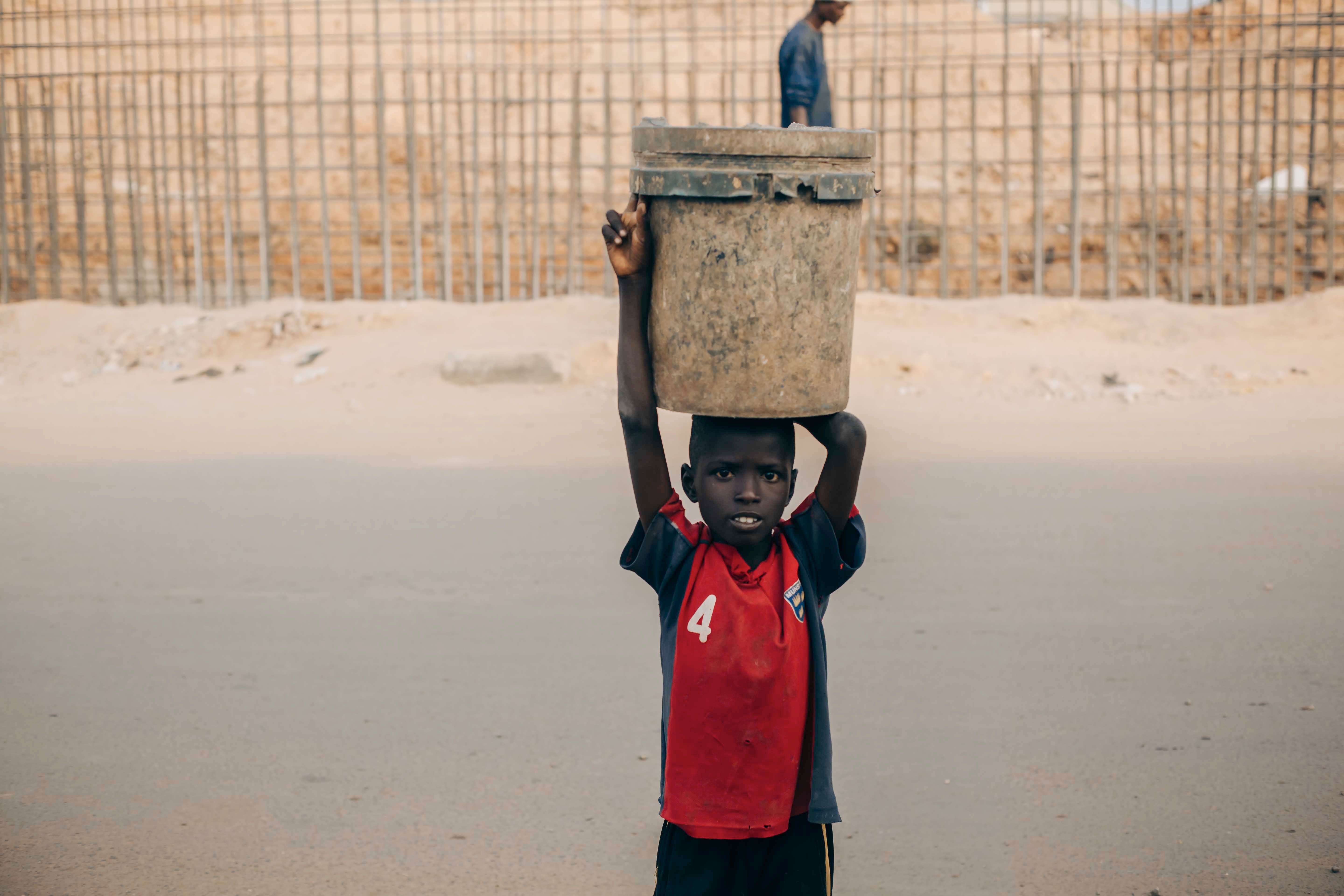Child carrying a large bucket on a street, embodying strength and resilience.