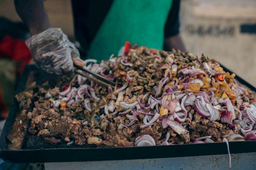 Chef prepares street food with chopped meat and onions on a hot griddle, using tongs.