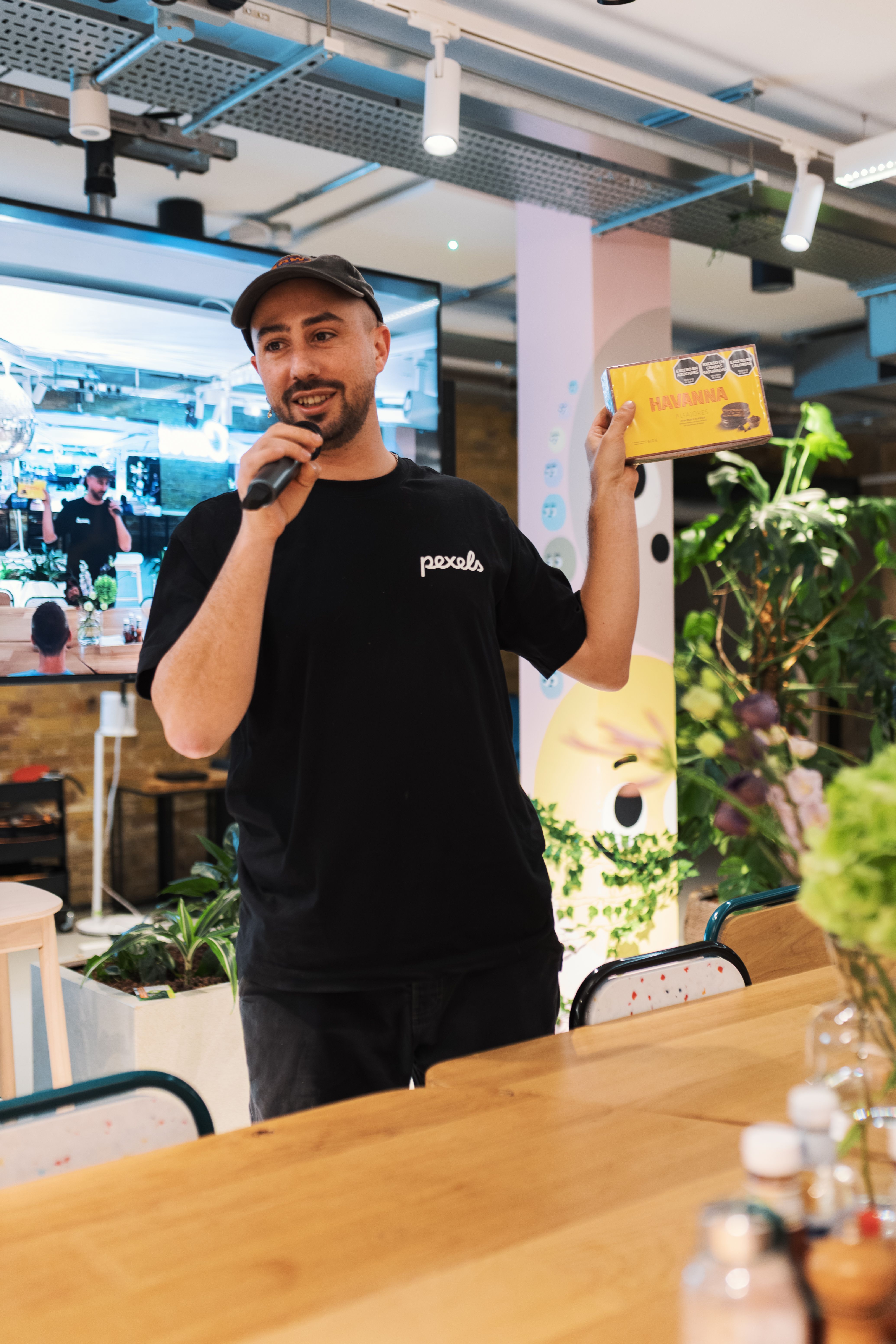A man in a black shirt giving a speech with a microphone at an indoor event with plants.