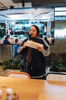 Woman holding microphone and paper, leading an indoor event with audience.