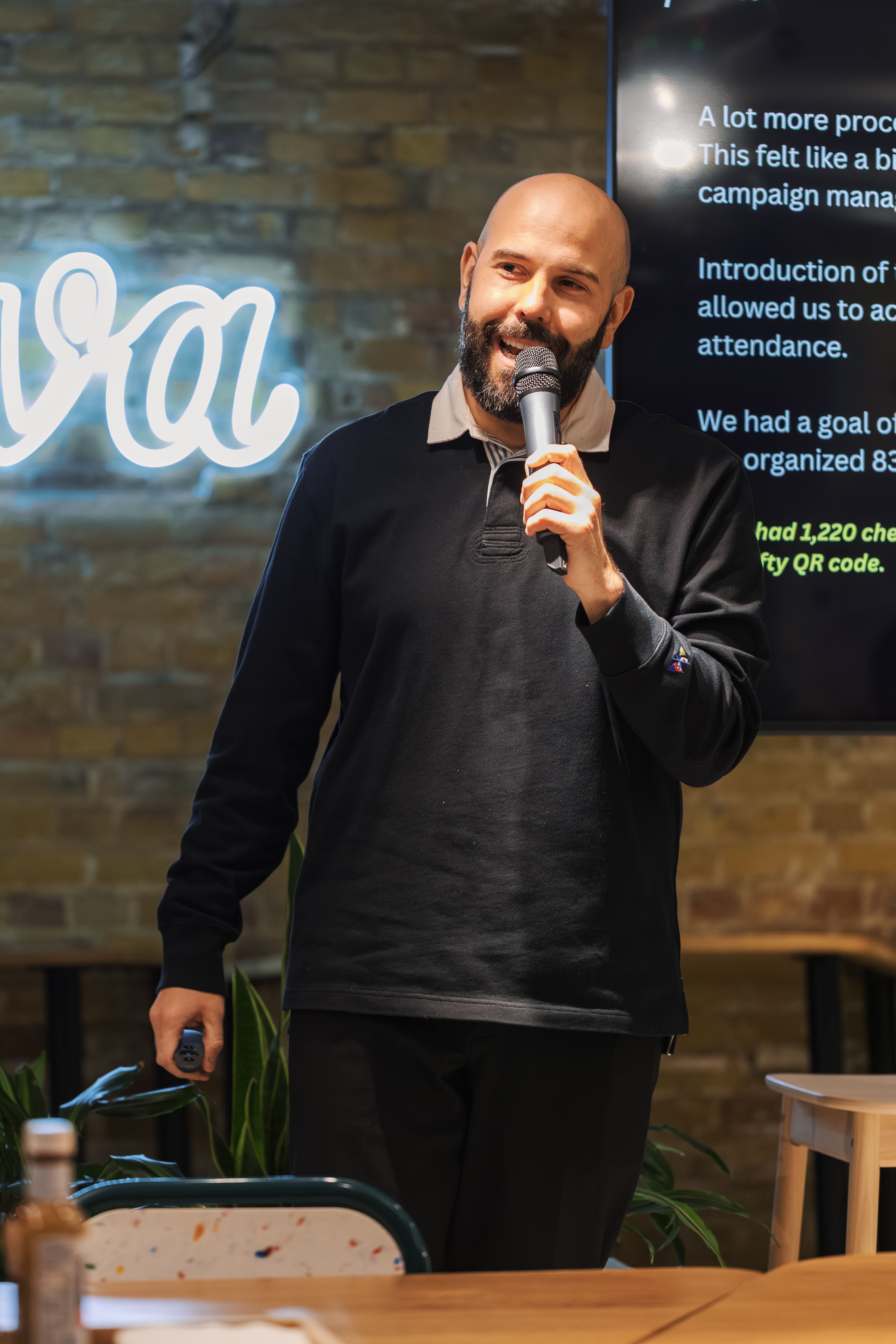 Bald man speaking with a microphone at an indoor event with rustic decor.