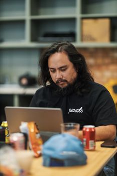 Man in a casual office setting working on a laptop with snacks and drinks nearby.