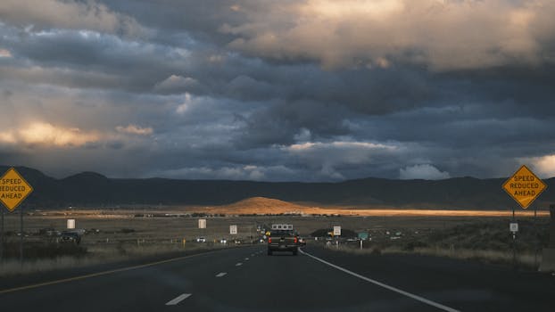 A car drives on a rural highway under dramatic sunset skies, surrounded by mountains.