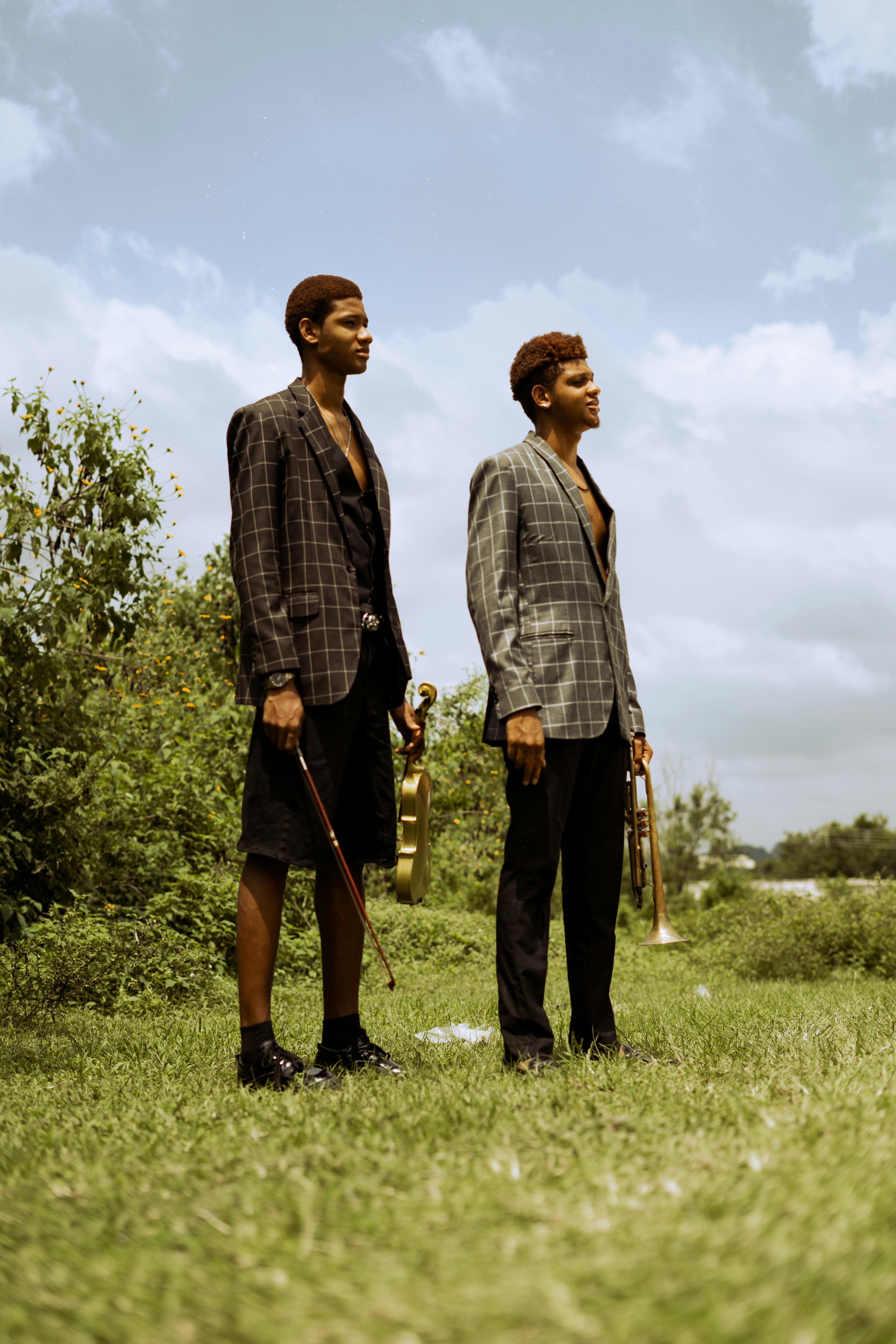 Two young men stand outdoors holding musical instruments, surrounded by greenery.