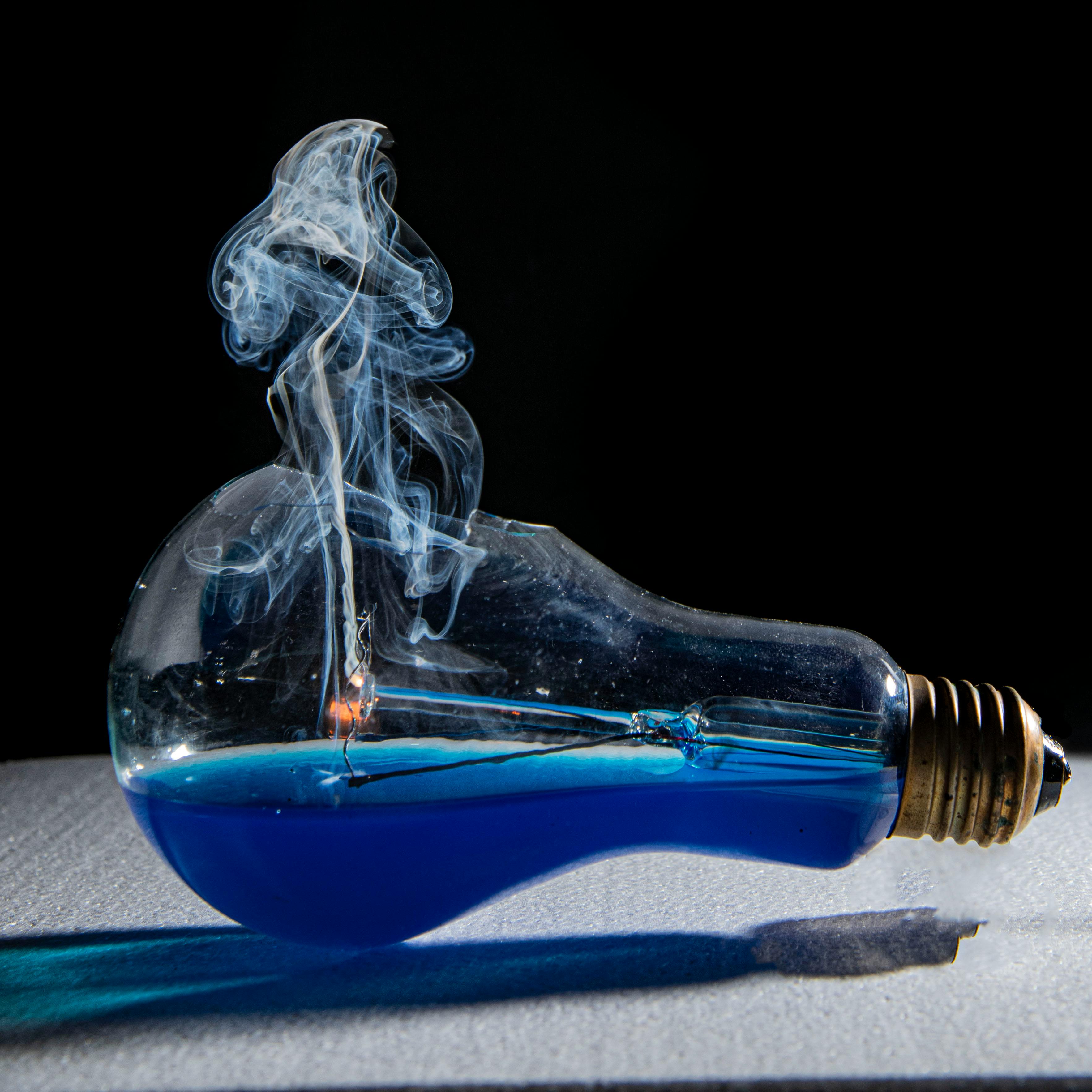 A creative shot of a lightbulb filled with blue liquid and artistic smoke rising, captured against a dark background.