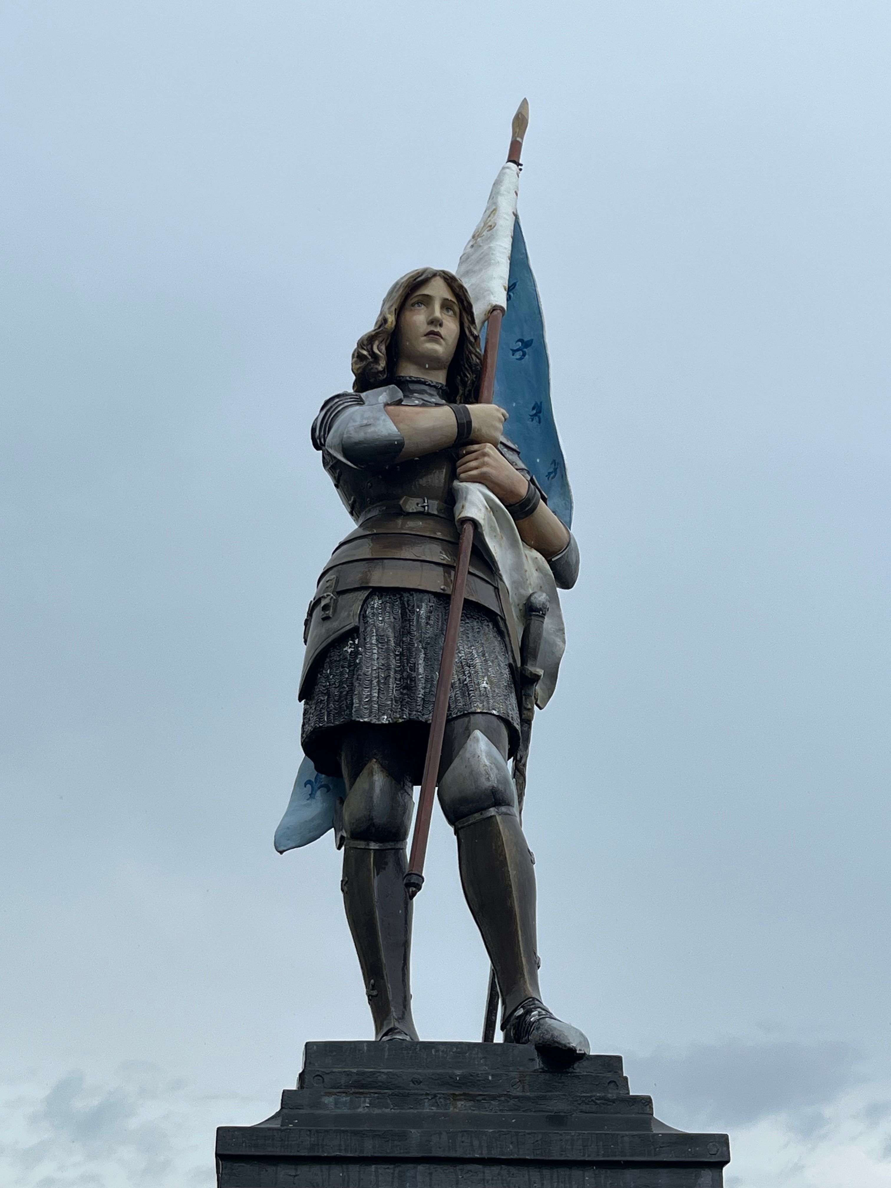 Majestic statue of a historic figure in armor holding a blue flag against a cloudy sky.