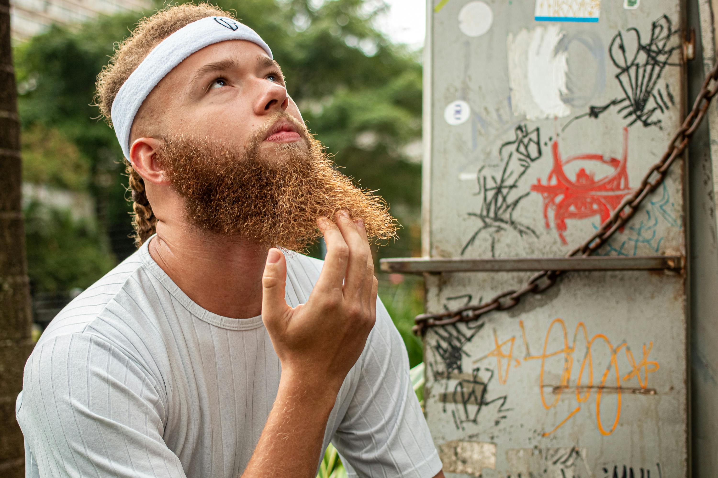 Bearded man in headband looking up thoughtfully outside near graffiti wall.