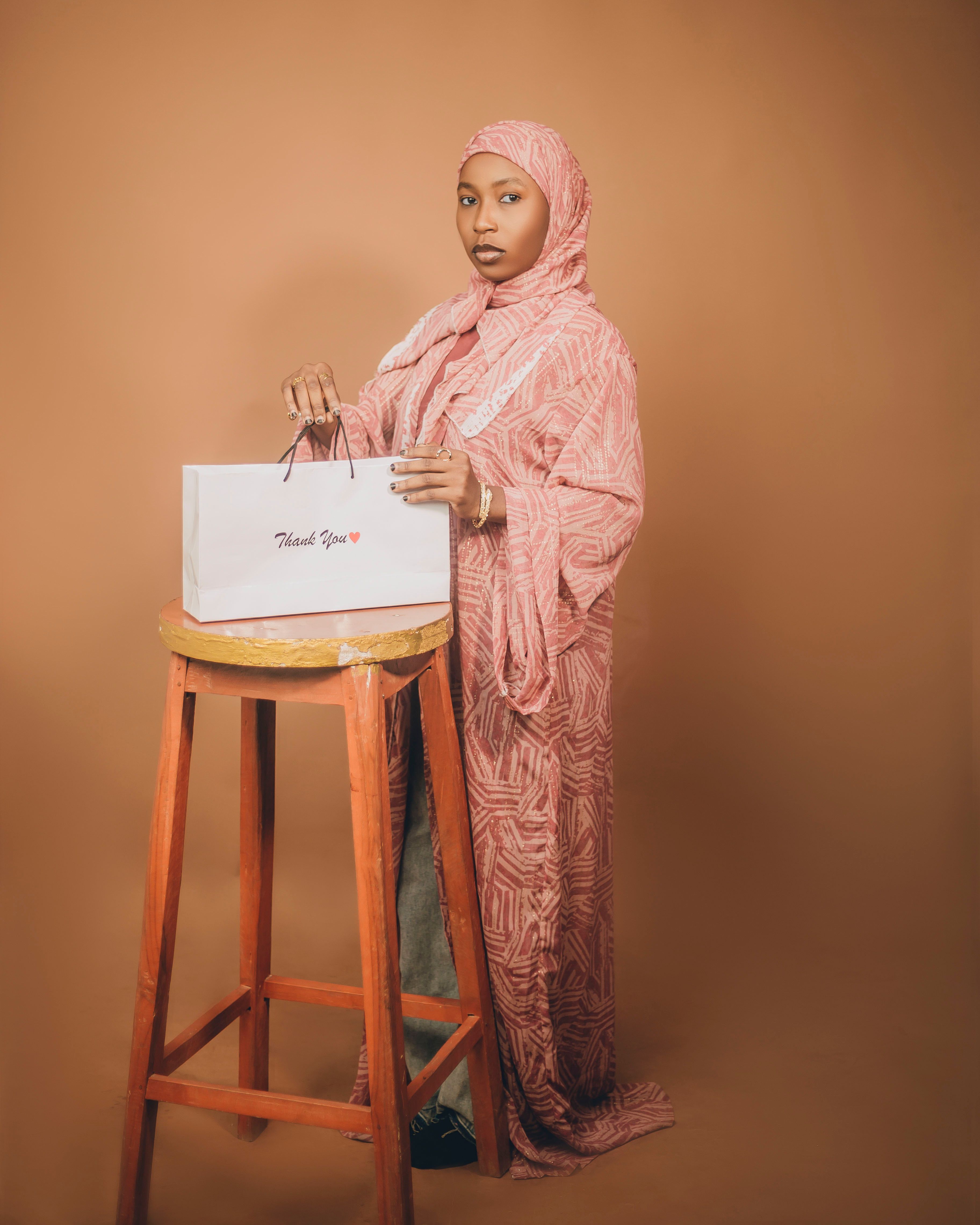 Young woman in a pink hijab holding a 'Thank You' bag indoors against a brown background.