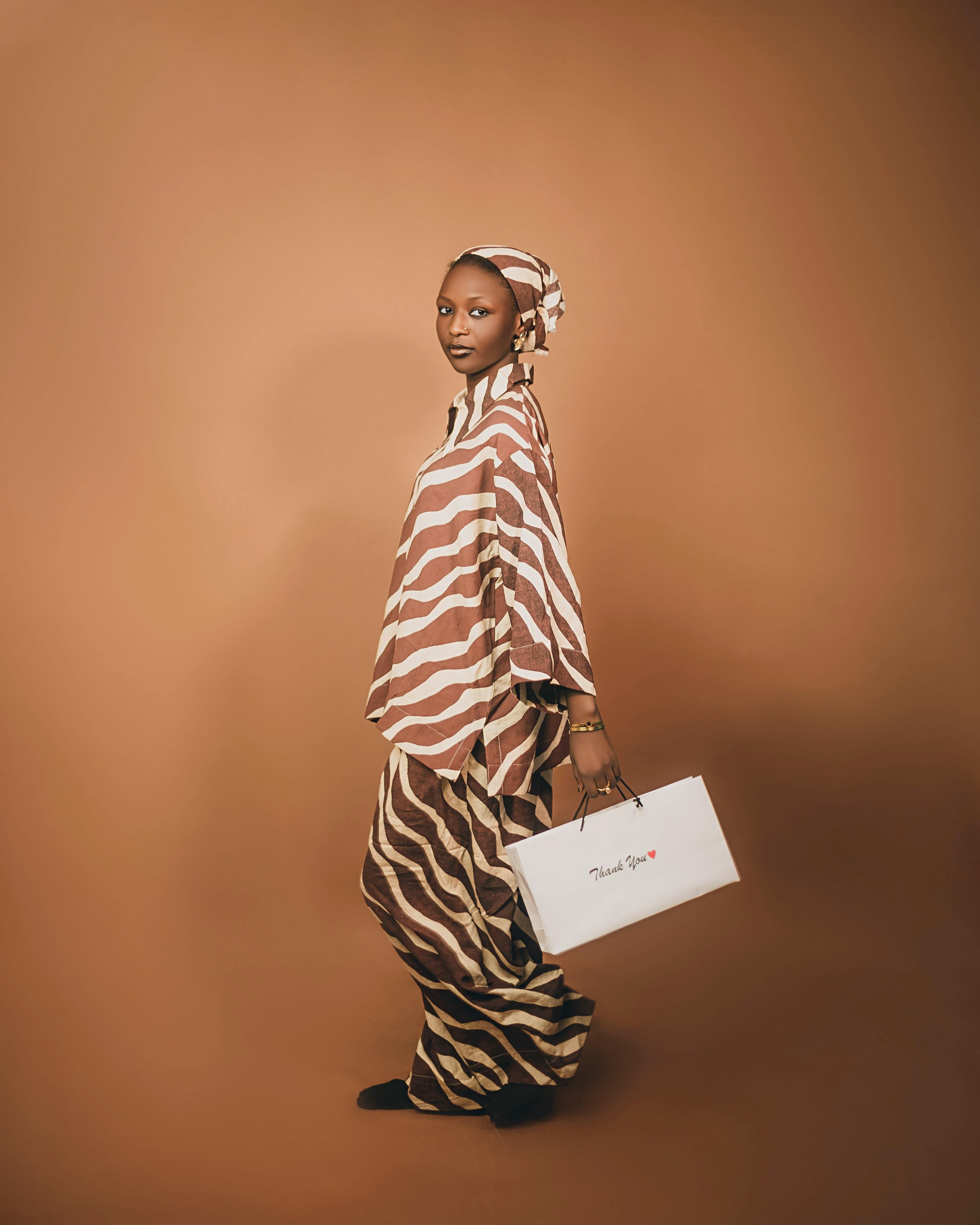 Elegant woman in fashionable traditional outfit with striped patterns and headscarf holding a bag.
