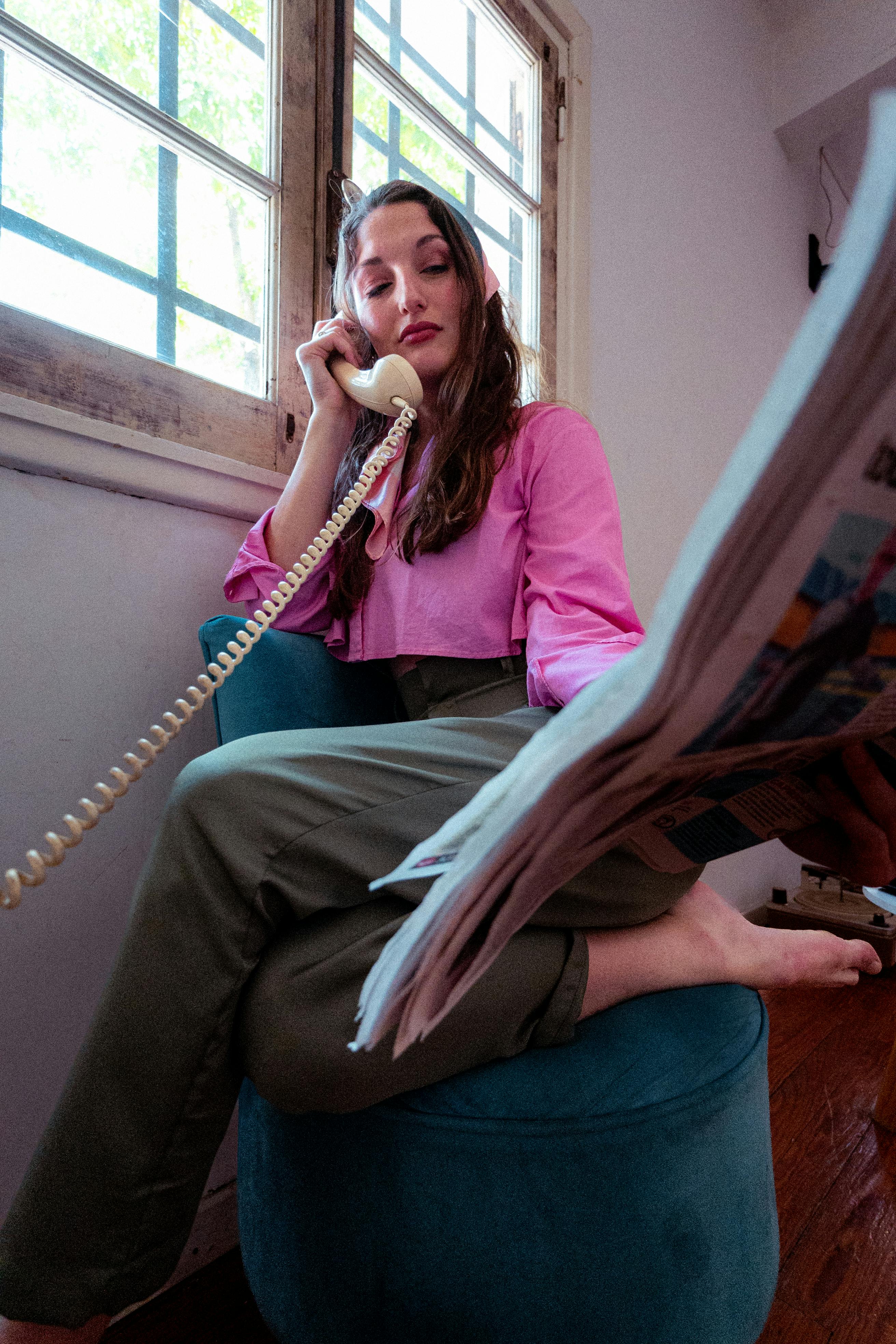 Ilmainen Nainen vintage-tyyliin lukee sanomalehteä ja käyttää pyöritettävää puhelinta ikkunan lähellä. Kuvapankkikuva