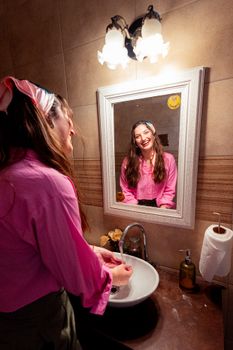 A joyful woman in a pink shirt smiles at her reflection in a vintage bathroom setting.