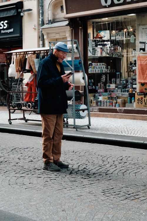Free An elderly man with a hat checks his smartphone on a cobblestone street beside quaint shops. Stock Photo