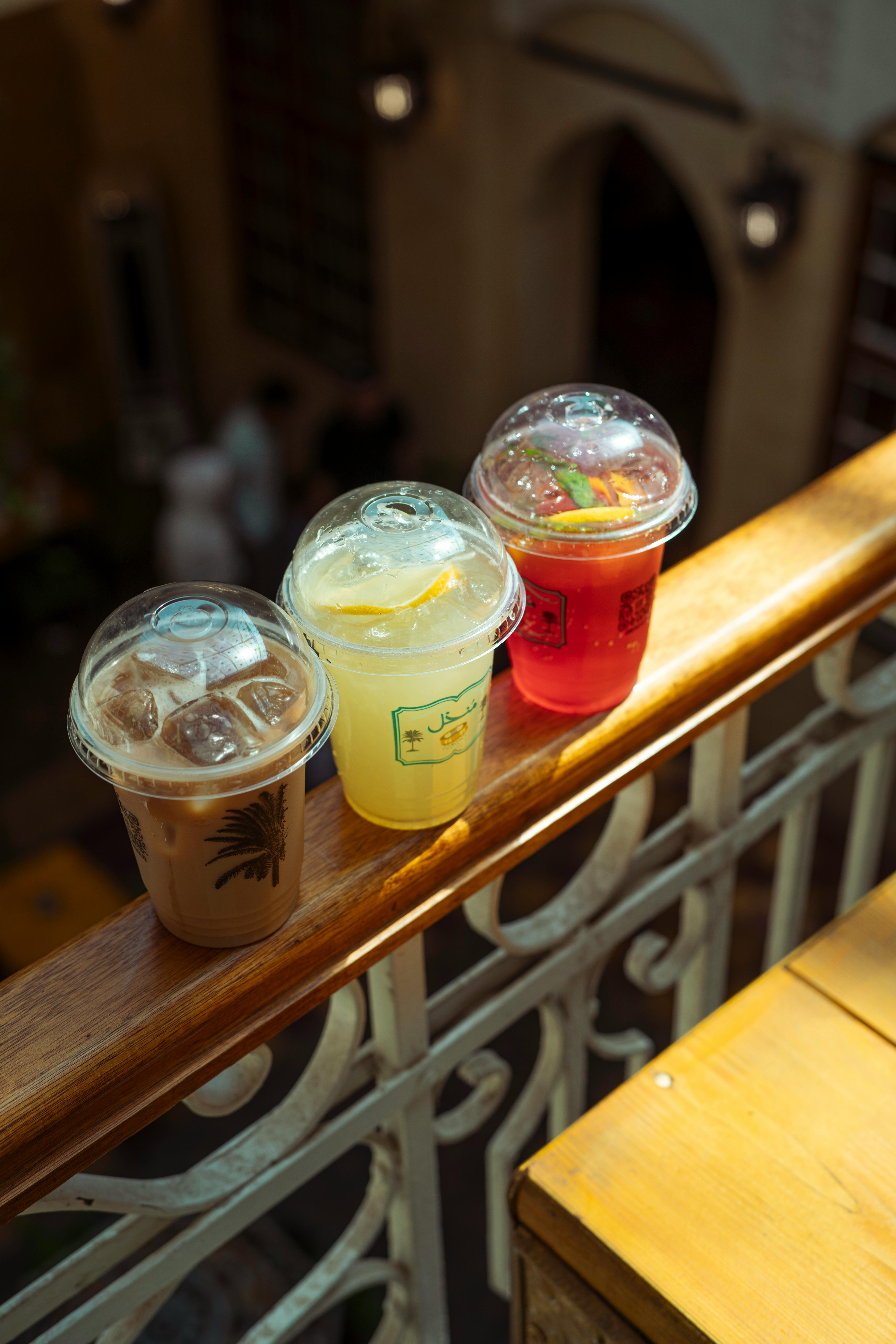 Three refreshing iced drinks sit on a sunlit balcony railing indoors.