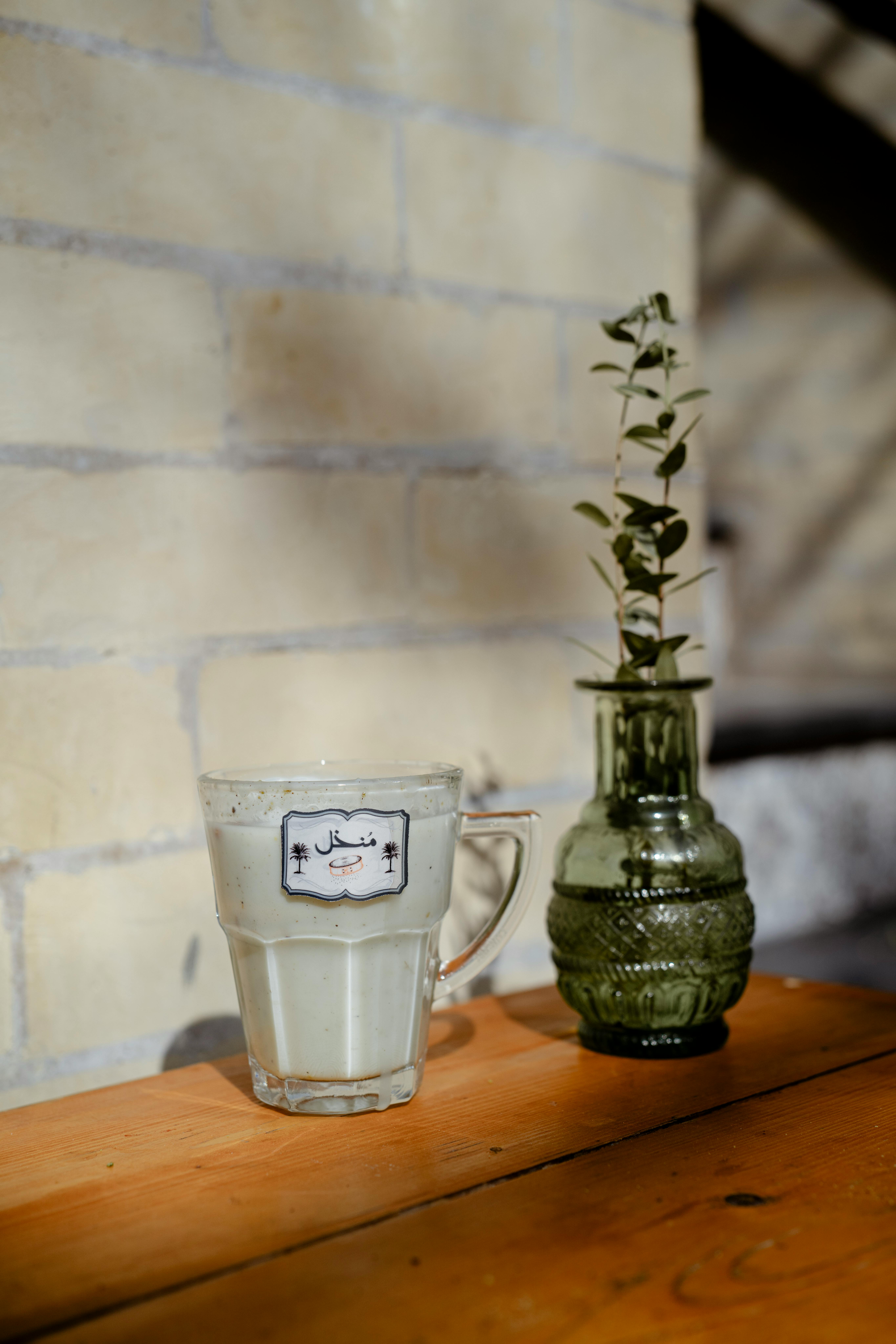 A glass of milk next to a decorative green vase on a wooden table casting shadows indoors.