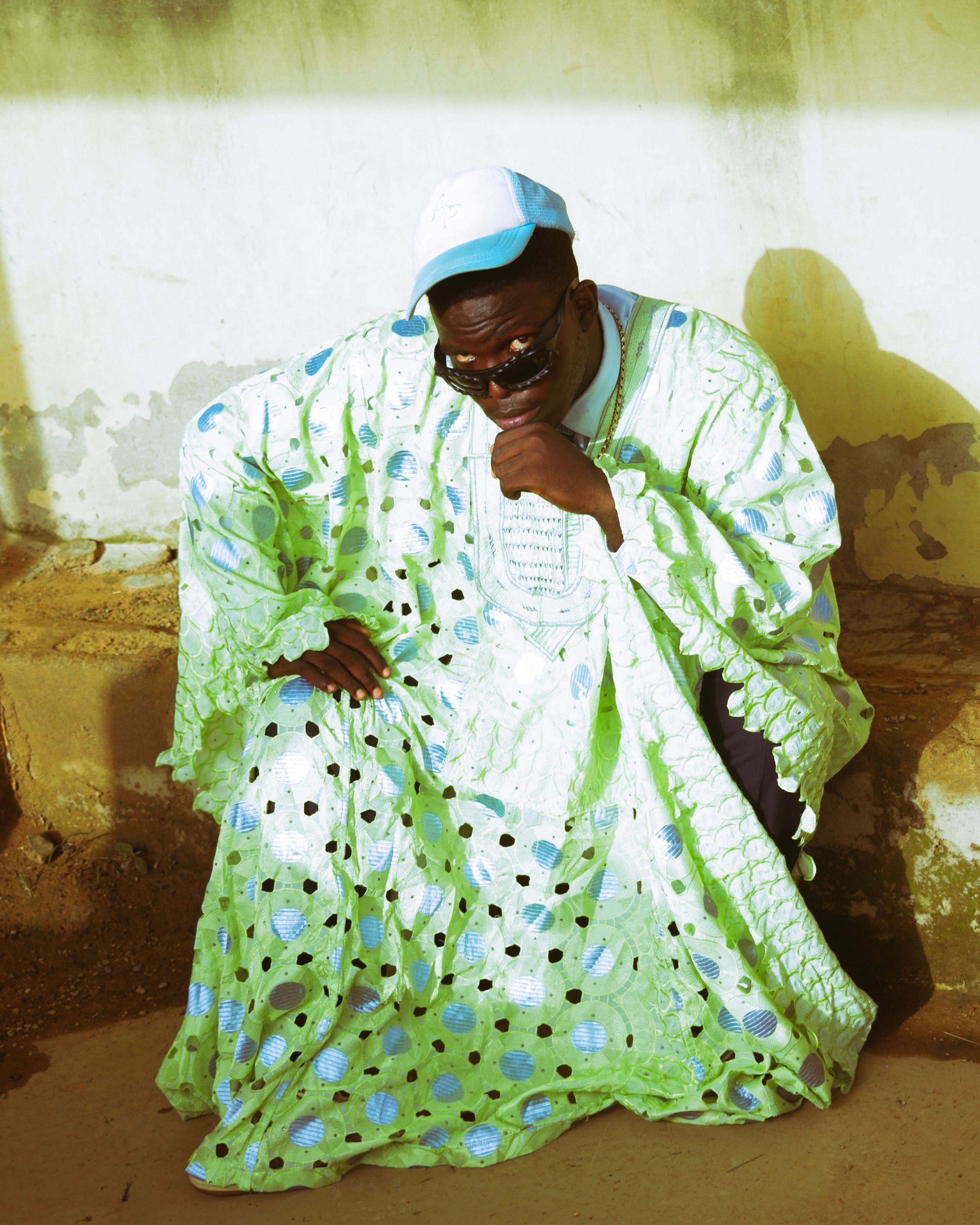 An adult man in vibrant traditional African clothing squatting outdoors.