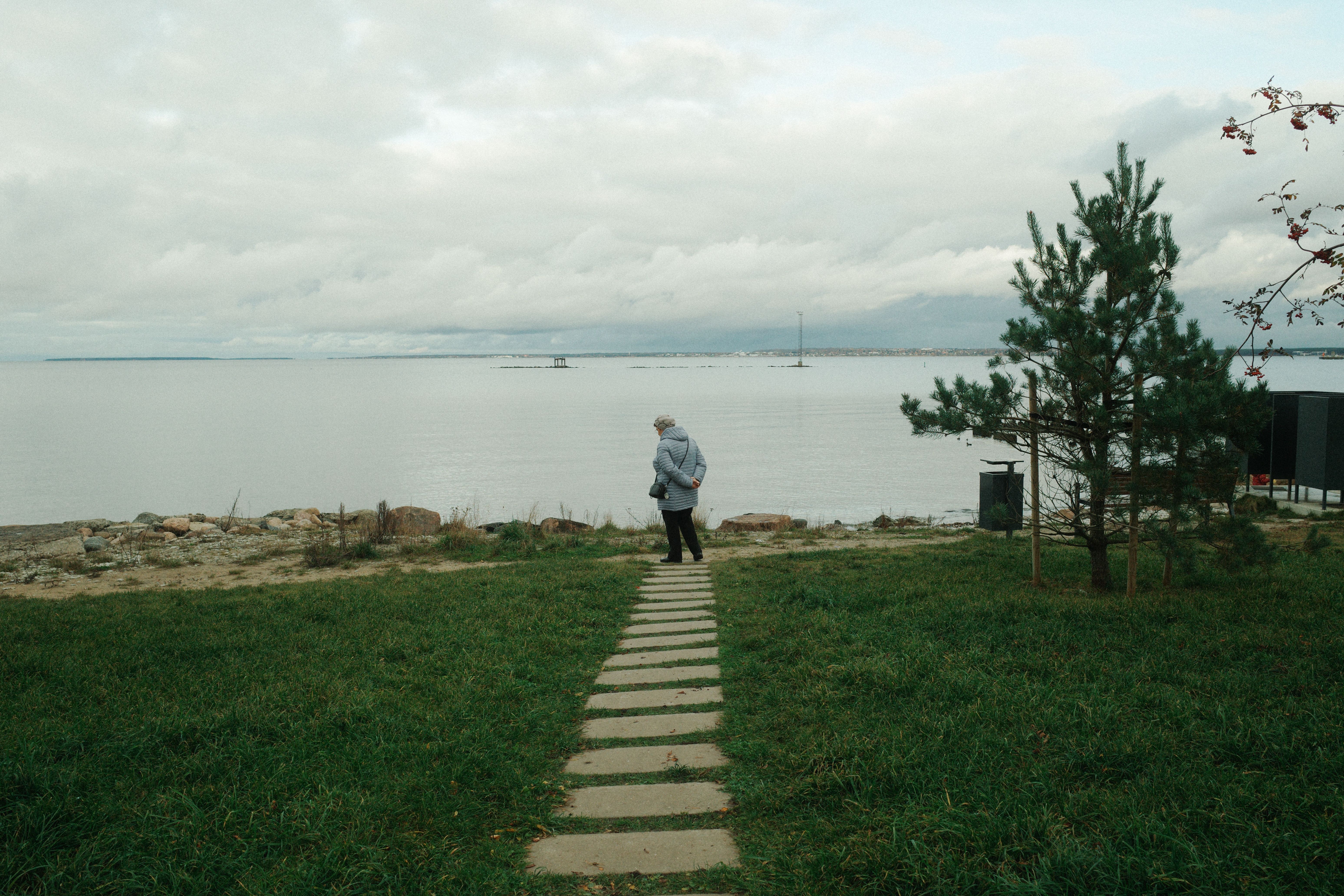 A person walks alone by a serene lake, surrounded by greenery and clouds.