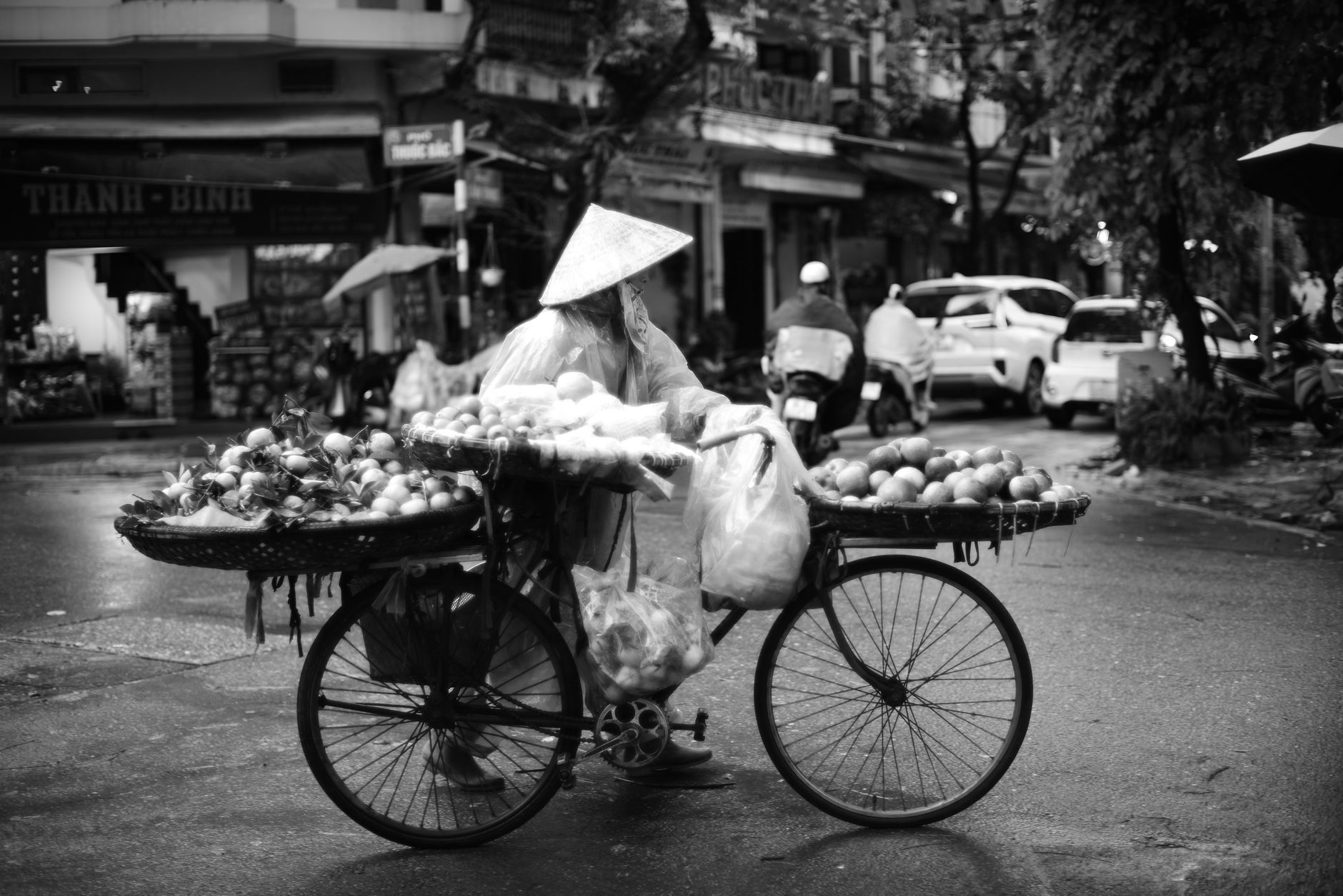 A street vendor with a traditional hat sells fruits from a bicycle in a bustling Vietnamese city.
