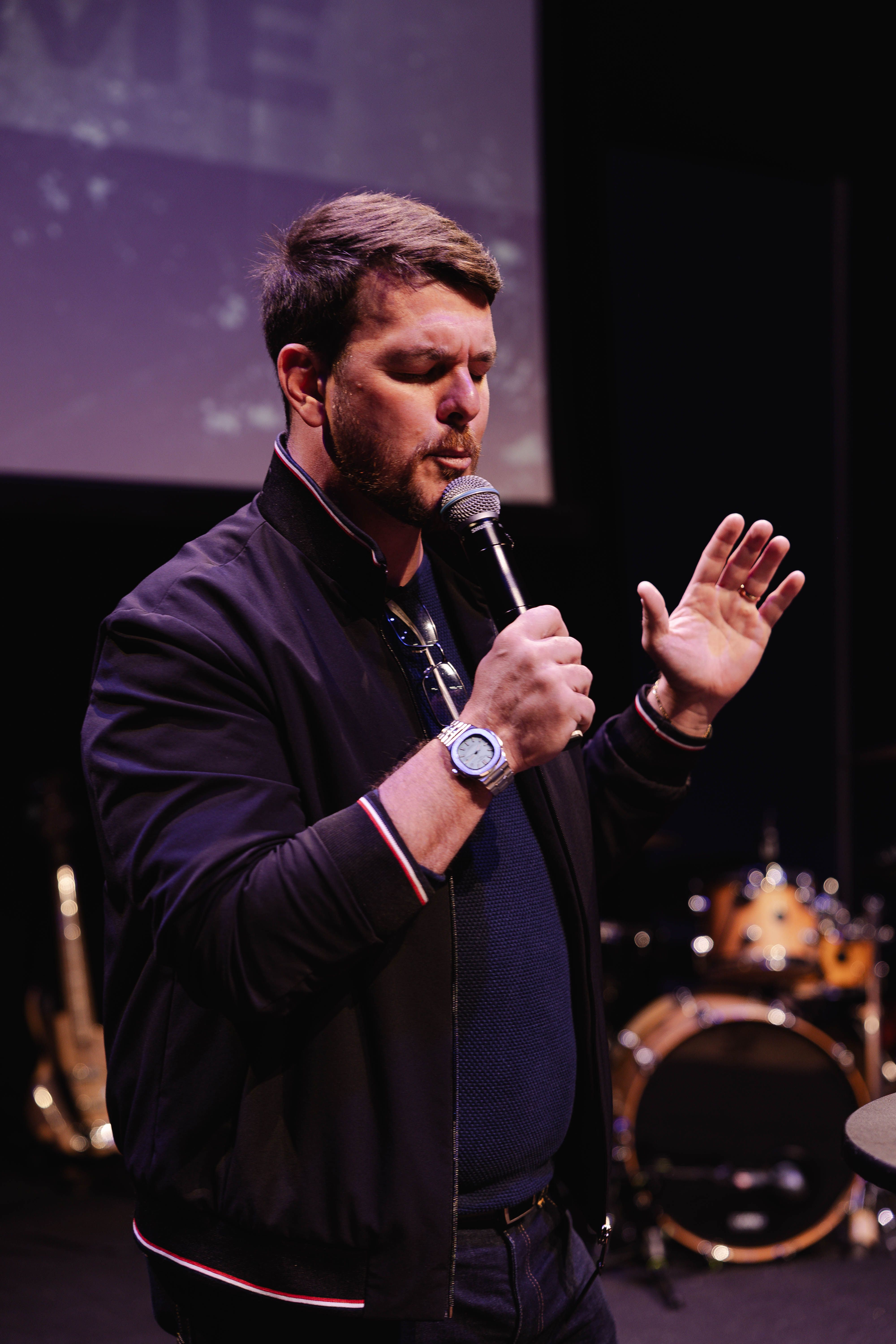 Adult male singing on stage with microphone, hands raised, engaging performance.
