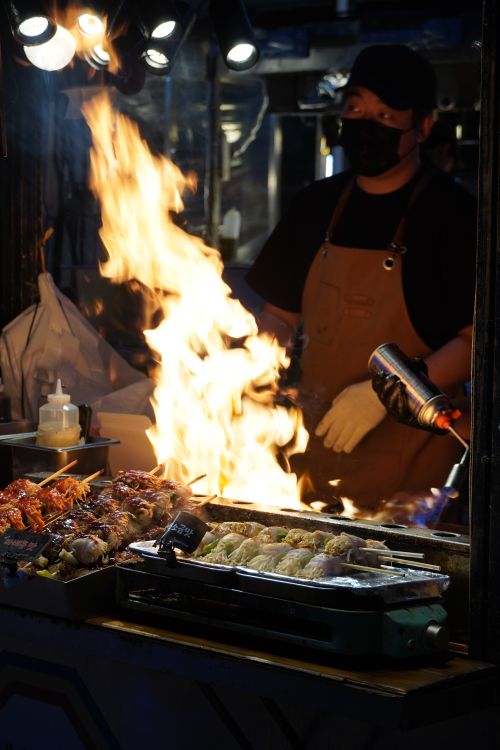 Free Vibrant night market scene of a street vendor grilling food with visible flames, capturing the lively atmosphere of South Korea. Stock Photo