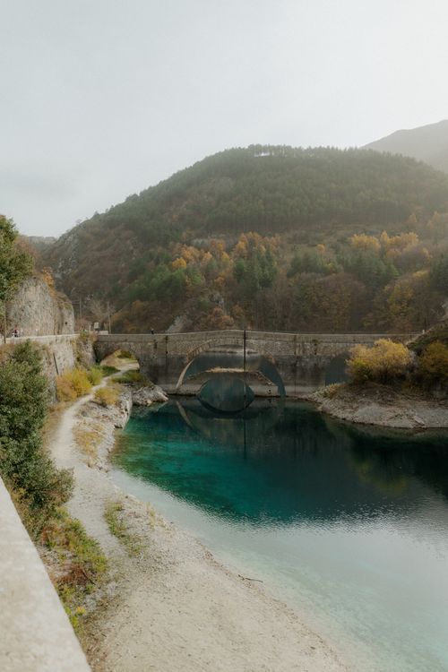 Free Beautiful stone bridge crossing a clear blue river surrounded by autumn foliage and mountains. Stock Photo