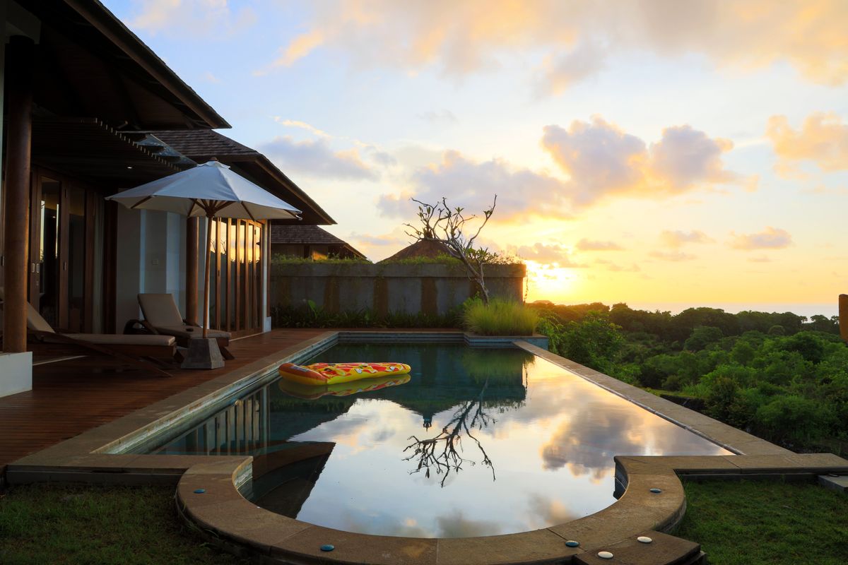 A serene view of a villa's infinity pool at sunset in Bali, Indonesia, with lush greenery and calm reflections.