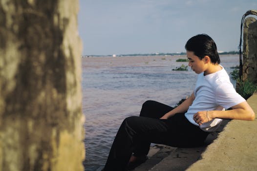 Young man in white shirt sitting riverside, reflecting quietly.