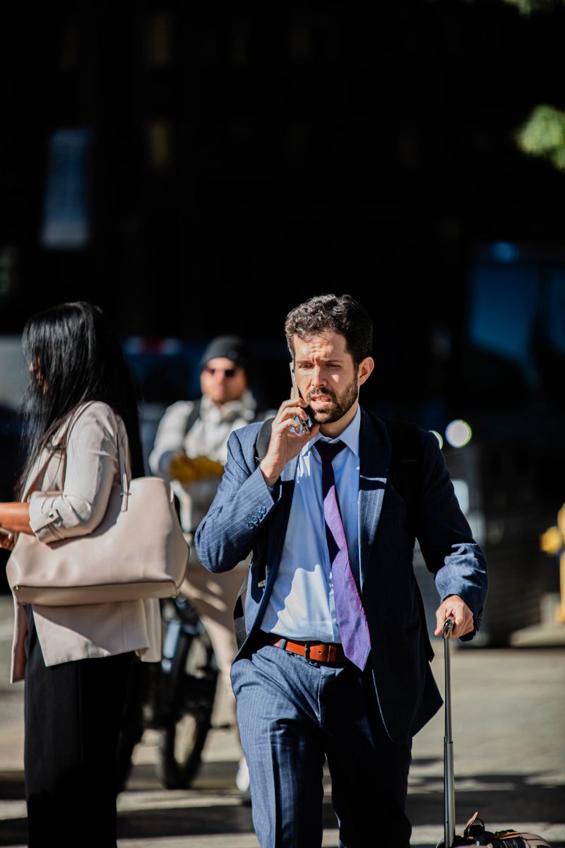 A businessman on a phone call walks with luggage on a city street