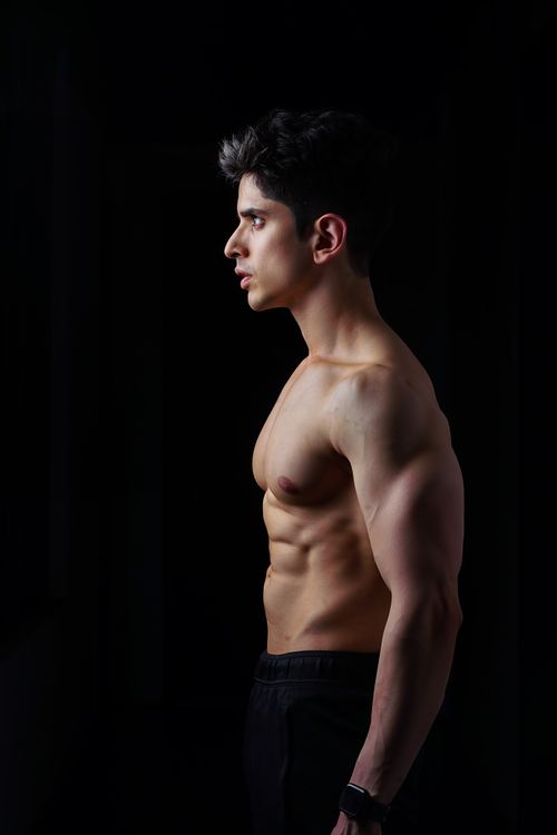 Free Side profile of a muscular young man standing in a dark studio setting, showcasing his physique. Stock Photo