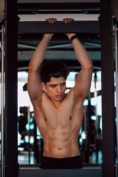 Muscular young man performing pull-ups on a bar in a modern gym setting.