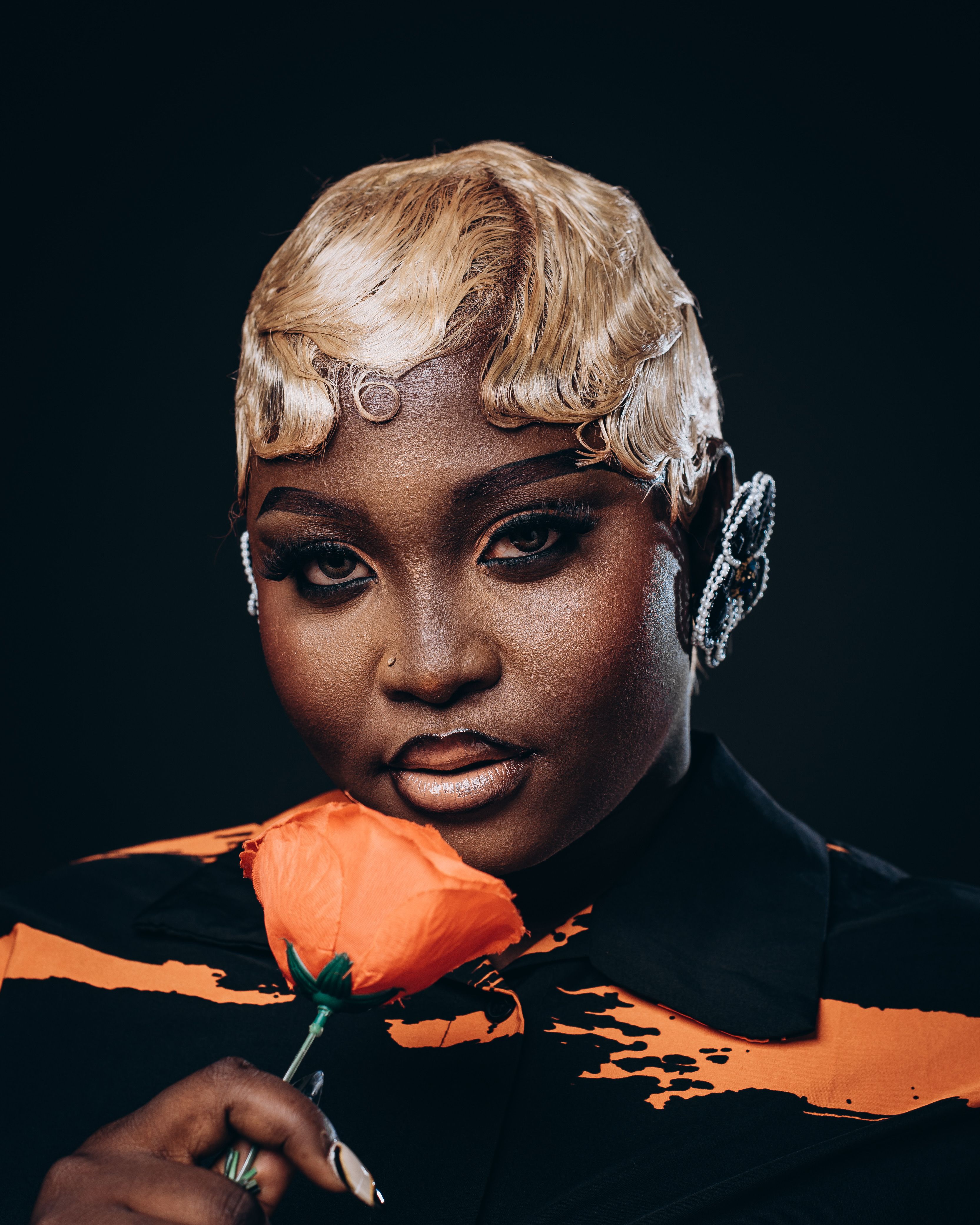 Elegant portrait of a woman with blonde hair holding a single orange flower against a dark background.