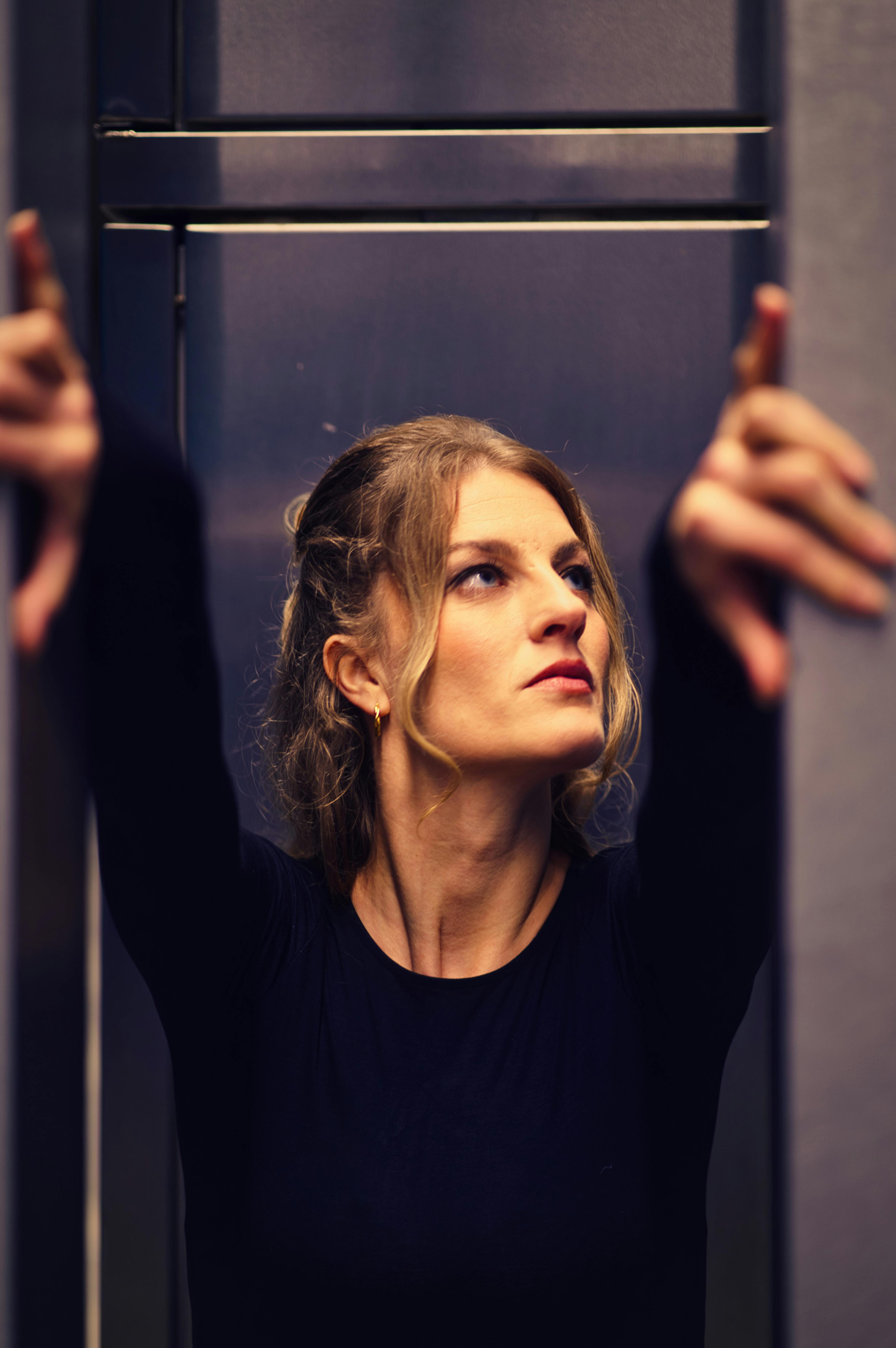 Woman with contemplative expression reaches upwards inside an elevator.