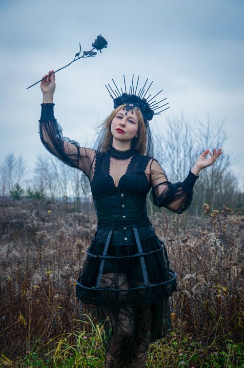 Free A woman in a gothic black dress poses dramatically with a flower in a moody outdoor setting. Stock Photo